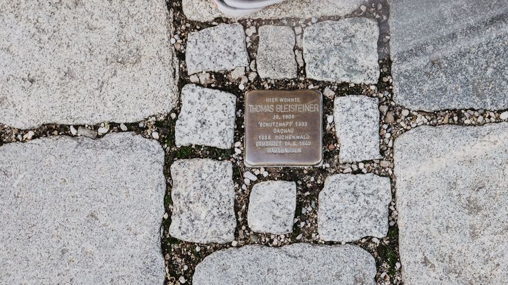 Ein Stolperstein im Pflaster der Dachauer Altstadt und eine Person, die daran vorbeigeht.